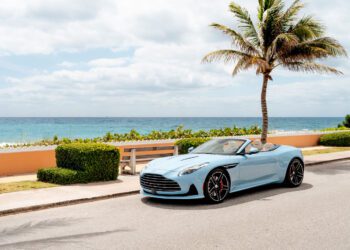 A light blue Aston Martin DB12 Volante convertible is parked on a coastal road near a palm tree, with the ocean and a partly cloudy sky in the background—capturing the essence of the exclusive Palm Beach Edition.