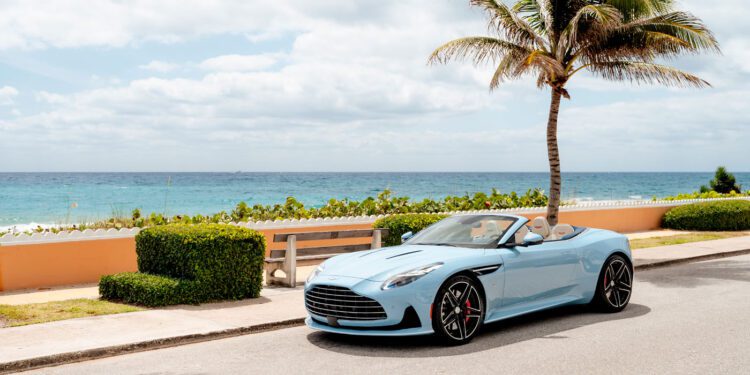 A light blue Aston Martin DB12 Volante convertible is parked on a coastal road near a palm tree, with the ocean and a partly cloudy sky in the background—capturing the essence of the exclusive Palm Beach Edition.