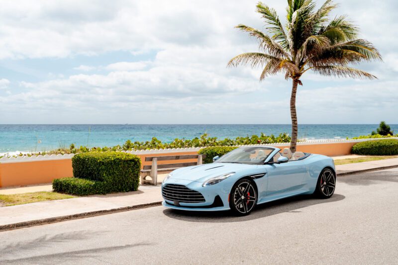 A light blue Aston Martin DB12 Volante convertible is parked on a coastal road near a palm tree, with the ocean and a partly cloudy sky in the background—capturing the essence of the exclusive Palm Beach Edition.
