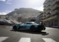 A teal and black Aston Martin sports car drives on a city street circuit, with tall buildings and a mountain visible in the background under a bright sky, evoking scenes from the Monaco Grand Prix.