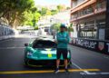 A person wearing a green jacket and cap stands with arms crossed in front of an Aston Martin sports car on a city street race track, echoing the thrilling energy of the Monaco Grand Prix.