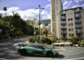 A green Aston Martin sports car drives around a sharp corner on a city street circuit lined with barriers, surrounded by tall buildings and greenery, reminiscent of the thrilling Monaco Grand Prix.