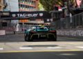 A green Aston Martin Valhalla speeds along a city racetrack near a TAG Heuer-branded finish line, evoking the excitement of the Monaco Grand Prix amid buildings and trees.