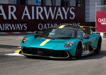A teal Aston Martin sports car with yellow accents drives on a racetrack, with Qatar Airways banners visible in the background, capturing the thrill of a Monaco Grand Prix moment.
