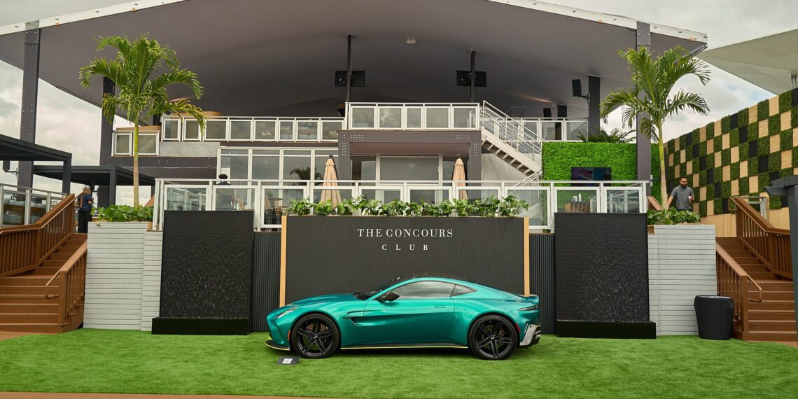 A turquoise sports car is parked on green artificial grass in front of a modern building labeled "The Concours Club," with palm trees and cloudy skies in the background.
