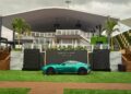 A turquoise sports car is parked on green artificial grass in front of a modern building labeled "The Concours Club," with palm trees and cloudy skies in the background.