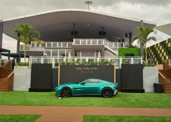 A turquoise sports car is parked on green artificial grass in front of a modern building labeled "The Concours Club," with palm trees and cloudy skies in the background.