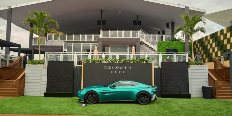 A turquoise sports car is parked on green artificial grass in front of a modern building labeled "The Concours Club," with palm trees and cloudy skies in the background.