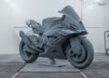 A gray, unfinished Carbon-Clad Concept BMW Motorrad full-scale motorcycle prototype stands on a pedestal in an industrial room, showcasing the next generation of RR design.