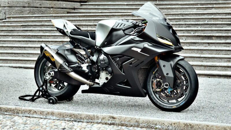 A matte black, carbon-clad concept BMW Motorrad showcases the next generation of RR as it sits on a gravel surface near concrete steps, supported by a rear wheel stand.