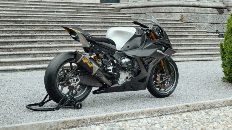 A modern Carbon-Clad Concept sport motorcycle with a black and white color scheme is parked on a gravel path in front of stone steps, showcasing BMW Motorrad’s next-generation RR design.