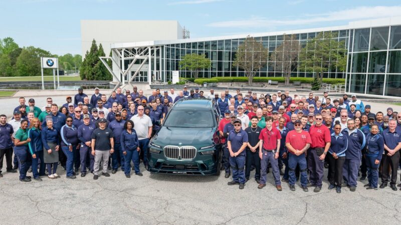 A large group of people poses around a green U.S.-Built BMW ALPINA XB7 SUV in front of a modern glass building on a clear day.