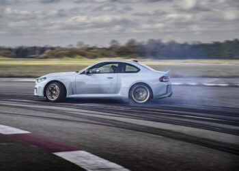 A white BMW 2026 M2 CS sports car drifting on a racetrack, with tire smoke and skid marks visible, under a cloudy sky—showcasing the power of its 523HP engine.