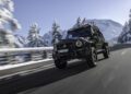 A Mercedes-AMG G63 drives on a snow-lined mountain road with snow-covered trees and peaks in the background under a clear sky.