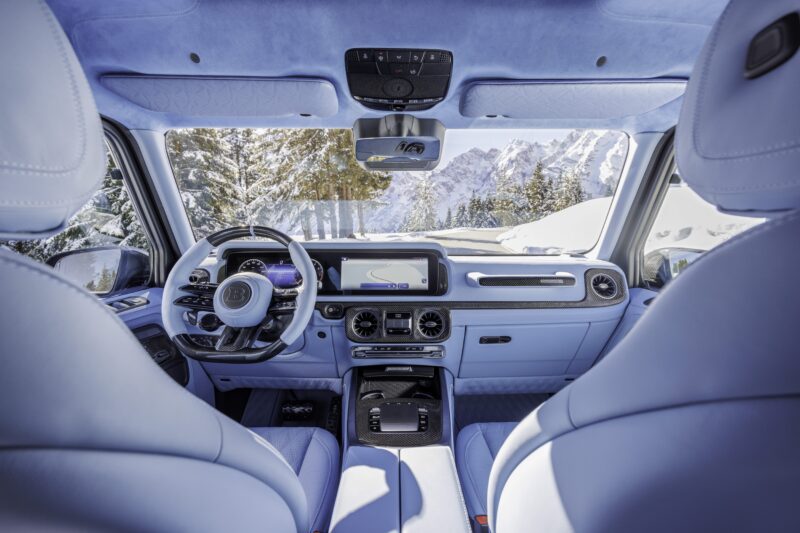 View from the back seat of a BRABUS XLP 800 ADVENTURE Mercedes-AMG G63 with blue leather interior, showcasing the dashboard, steering wheel, and a snowy mountain landscape through the windows.