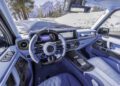 Steering wheel and dashboard of a BRABUS XLP 800 ADVENTURE Mercedes-AMG G63 with blue leather interior, digital displays, and carbon fiber accents, parked by a snowy mountain road.
