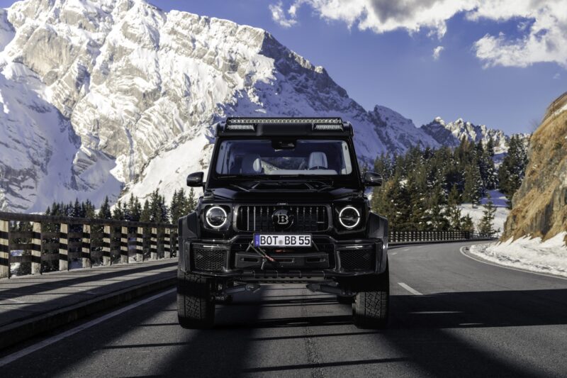 A black BRABUS XLP 800 ADVENTURE, based on the Mercedes-AMG G63, is parked on a mountain road with snowy peaks and pine trees in the background under a partly cloudy sky.