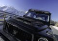 A black BRABUS XLP 800 ADVENTURE, based on the Mercedes-AMG G63, stands parked on a mountain road, its carbon fiber hood gleaming against snow-covered peaks and a wooden guardrail beneath the clear sky.