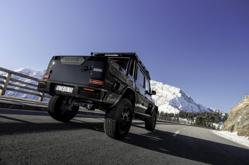 A BRABUS XLP 800 ADVENTURE, based on the Mercedes-AMG G63, with off-road modifications drives on a paved mountain road against snow-covered peaks and a clear blue sky.