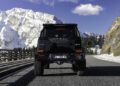 A BRABUS XLP 800 ADVENTURE black SUV drives on a mountain road with snow-covered peaks and trees in the background under a clear blue sky.