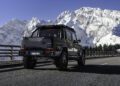 A BRABUS XLP 800 ADVENTURE off-road vehicle drives on a paved mountain road, framed by snow-covered peaks and a clear blue sky in the background.