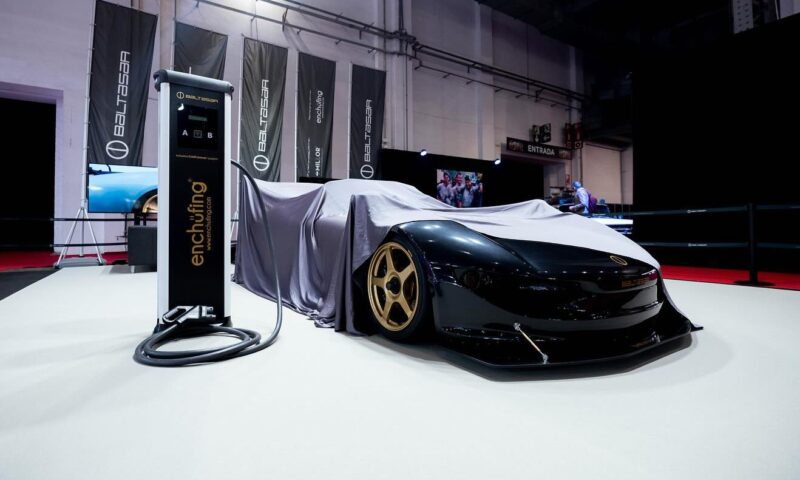 A sleek black sports car with gold rims and FIA cage is partially covered by a grey cloth at an indoor auto show, next to an electric vehicle charging station—could this be the new king EV of track days?.