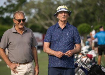 Two older men stand on a golf course, one wearing sunglasses and a polo shirt, the other in a patterned shirt and bucket hat. Golf bags and people are visible in the background.
