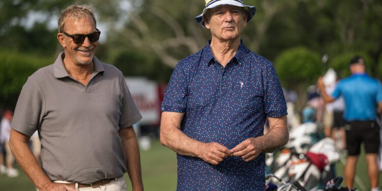 Two older men stand on a golf course, one wearing sunglasses and a polo shirt, the other in a patterned shirt and bucket hat. Golf bags and people are visible in the background.