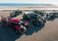Five vintage open-top cars, including a Blower Jnr, are parked on a paved area by a beach with the ocean in the background under a clear sky, ready for production by Bentley & Hedley Studios.