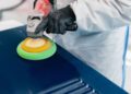 A person at Bentley & Hedley Studios, wearing gloves and a protective suit, uses a power buffer with a green and yellow pad to polish a blue surface during production.