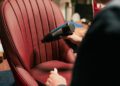 A person uses a Blower Jnr heat gun on a red leather chair in the workshop at Bentley & Hedley Studios, likely for upholstery or repair work as part of the production process.