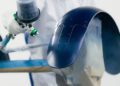A person in protective clothing at Bentley & Hedley Studios spray paints a curved metal part with dark blue paint in a controlled production environment.