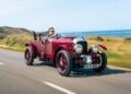A man drives a vintage red Blower Jnr convertible, crafted by Bentley & Hedley Studios, along a coastal road with the ocean and hills in the background.