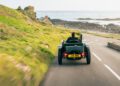 A person drives a vintage Blower Jnr convertible along a winding coastal road bordered by grass and rocks, with the sea visible in the background—a classic scene reminiscent of Bentley & Hedley Studios’ timeless production style.