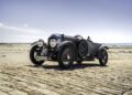 A vintage blue Bentley Blower Jnr open-top car with large headlights is parked on a cobblestone surface by a beach under a clear sky, capturing Hedley Studios’ signature classic style.