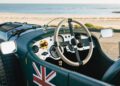 Close-up view of a vintage Bentley’s steering wheel and dashboard, with a British flag on the door, parked by the beach with the ocean in the background. A true Hedley Studios classic from the Blower Jnr Production collection.