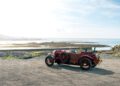 A vintage red Bentley Blower Jnr convertible by Hedley Studios is parked on a gravel area near a rocky shore, with the sea and a cloudy sky in the background.