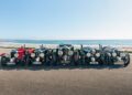 Six vintage cars, including a Bentley Blower Jnr, are parked side by side on a paved road with the ocean and a clear sky in the background—a stunning scene reminiscent of Hedley Studios' timeless style.