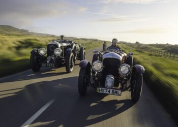 Two vintage cars, including a Blower Jnr, with drivers travel on a scenic road through the countryside. A blurred background suggests motion on this Bentley & Hedley Studios production, set against clear skies above.