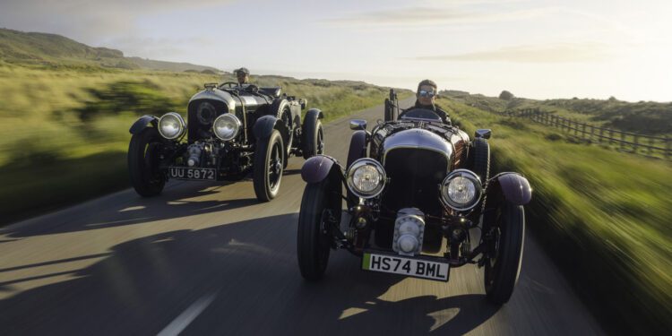 Two vintage cars, including a Blower Jnr, with drivers travel on a scenic road through the countryside. A blurred background suggests motion on this Bentley & Hedley Studios production, set against clear skies above.