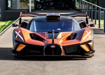 A front view of an orange and black $4.7Million Bugatti Bolide with copper carbon fiber accents and its doors open, parked on pavement near a modern building.