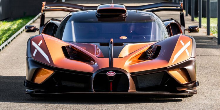 A front view of an orange and black $4.7Million Bugatti Bolide with copper carbon fiber accents and its doors open, parked on pavement near a modern building.