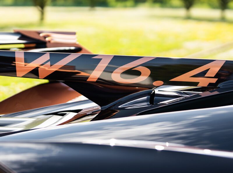 Close-up of a Bugatti Bolide's rear wing crafted from carbon fiber, featuring "W 16.4" in bold orange letters, set against a blurred outdoor background.