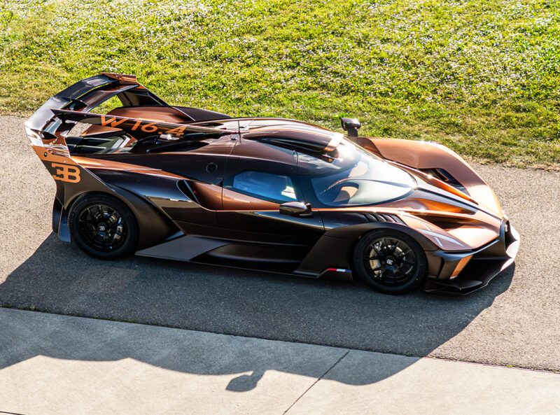 A black and copper Bugatti Bolide track car with carbon fiber aerodynamic features is parked on a paved surface beside grass, showcasing its $4.7Million exclusivity.
