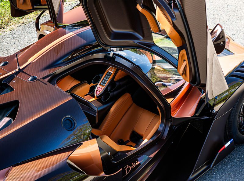A $4.7Million Bugatti Bolide with its doors open, featuring a tan leather interior, modern dashboard, and striking copper carbon fiber exterior.