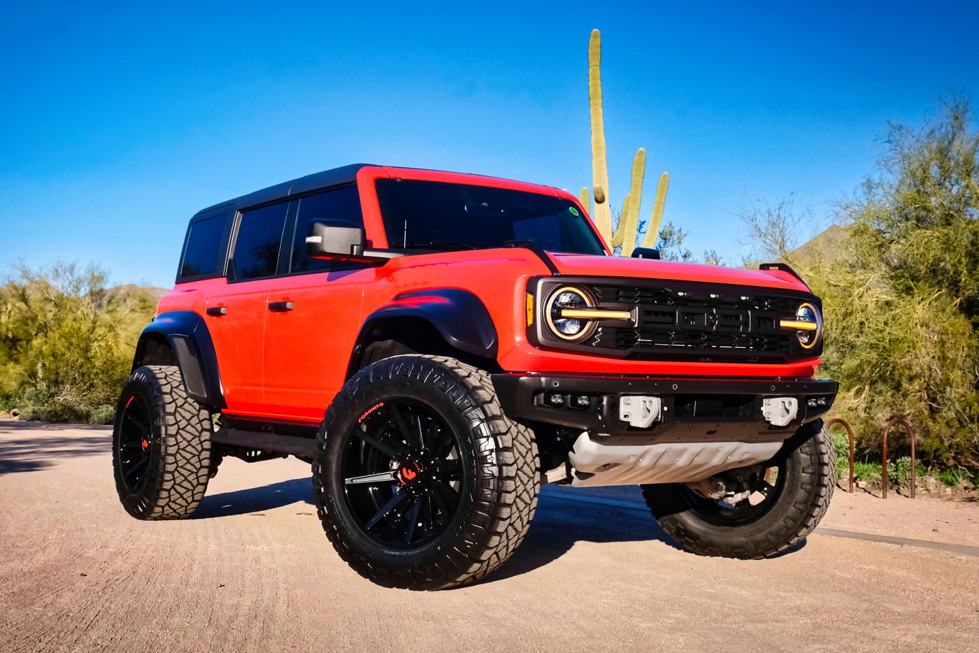 A red Ford Bronco SUV with black accents and large off-road tires is parked on a desert road, surrounded by cacti and desert plants under a clear blue sky—a perfect example from America’s Original Lifted Truck Dealer.
