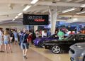 People attend an indoor car auction event with classic cars on display and a large "Carlisle Auctions" sign hanging from the ceiling at the 2025 Spring Carlisle Auction.