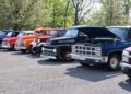 A row of vintage trucks and cars parked outdoors at a car show, with one truck's hood open; trees and greenery in the background hint at the excitement of the upcoming 2025 Spring Carlisle Auction by Carlisle Auctions.