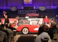 A red and white classic convertible is on display at the 2025 Spring Carlisle Auction, with auctioneers and attendees watching and bidding.