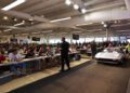A crowd sits at long tables watching an auctioneer as a silver sports car is driven onstage at the 2025 Spring Carlisle Auction, capturing the excitement of a Carlisle Auctions event.